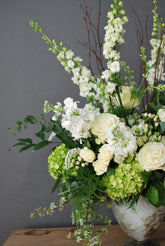 Side shot of a sympathy floral arrangement in green and whites, in a large vessel placed on a wooden table in front of a grey background. 