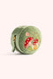 A small round sage-green velvet jewelry box with a zipper closure, featuring embroidered mushrooms and flowers on the front, set against a solid pale pink background.