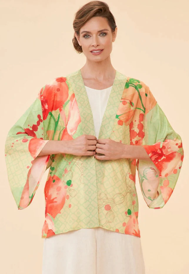 A woman wearing a light green kimono with a vibrant coral-and-orange floral print, layered over a white top and white pants, set against a solid beige background.