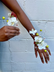 Hands modeling a corsage and boutonniere in whites and greens against a white wall. Corsage and boutonniere are both on wire bases.
