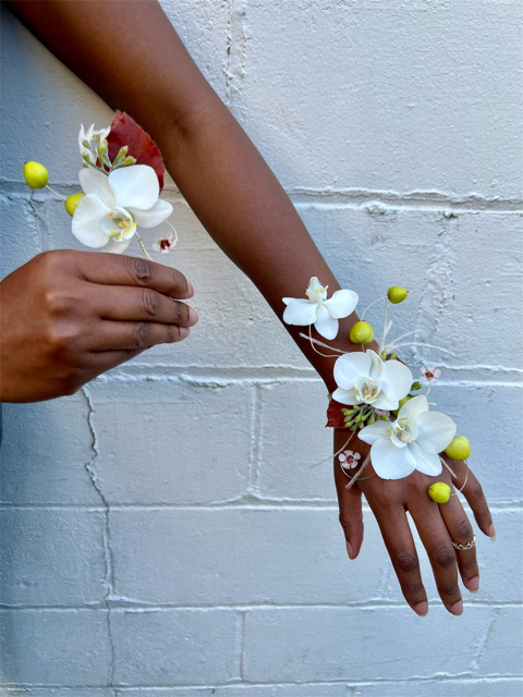 Hands modeling a corsage and boutonniere in whites and greens against a white wall. Corsage and boutonniere are both on wire bases.
