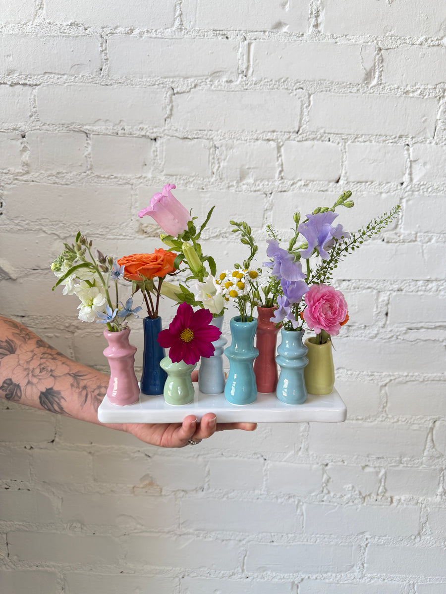 Eight colorful budvases set on a ceramic base, filled with colorful flowers in a wildflower style, against a white brick background. 