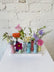 Eight colorful budvases set on a ceramic base, filled with colorful flowers in a wildflower style, against a white brick background. 
