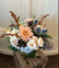 A floral arrangement made in a neutral fall color palette with dried elements popping out of the top, sitting on a wooden crate in front of a white background.