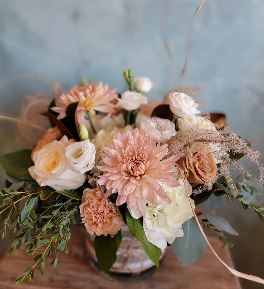 Floral arrangement in a neutral pink and white color palette with whispy greens and accent, in a vase wrapped in a burlap inspired ribbon.