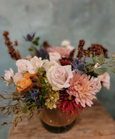 A floral arrangement made in a traditional muted autumn color palette, in a vase with a pumpkin-orange ribbon wrapped around the vase.