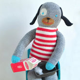 A handmade knitted dog doll with a gray body, red and white striped sweater, blue feet, and a brown face, sitting on a teal chair and holding a miniature milk carton.