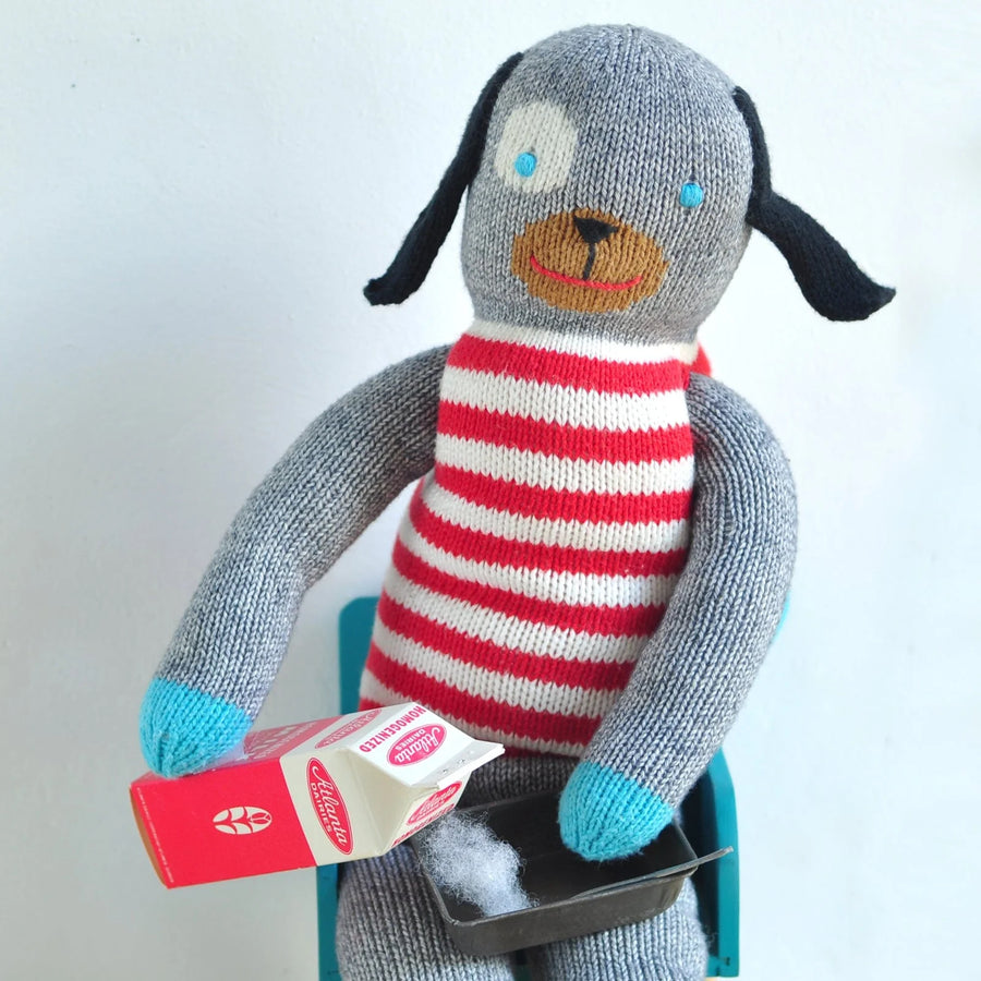 A handmade knitted dog doll with a gray body, red and white striped sweater, blue feet, and a brown face, sitting on a teal chair and holding a miniature milk carton.