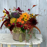 Floral arrangement in fall colors including yellow pincushion protea, black eyed susans, kale, and orange dahlias, in a round vase with burlap wrapped around it. The arrangement sits on a white wooden side table, in front of a grey washed wooden door. 
