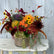 Floral arrangement in fall colors including yellow pincushion protea, black eyed susans, kale, and orange dahlias, in a round vase with burlap wrapped around it. The arrangement sits on a white wooden side table, in front of a grey washed wooden door. 
