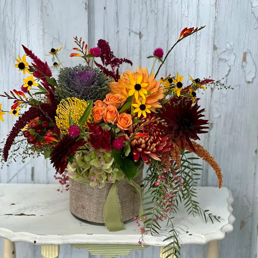 Floral arrangement in fall colors including yellow pincushion protea, black eyed susans, kale, and orange dahlias, in a round vase with burlap wrapped around it. The arrangement sits on a white wooden side table, in front of a grey washed wooden door. 
