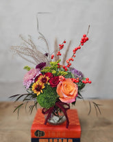 A small fall colored floral arrangement in a clear round vase. The arrangement sits atop a stack of books on a wooden table.