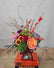 A small fall colored floral arrangement in a clear round vase. The arrangement sits atop a stack of books on a wooden table.