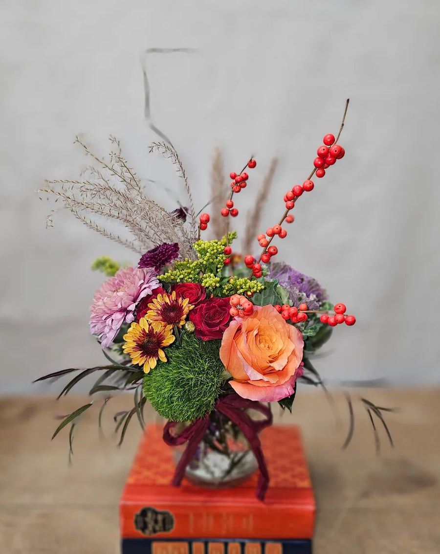 A small fall colored floral arrangement in a clear round vase. The arrangement sits atop a stack of books on a wooden table.