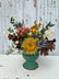 Small floral arrangement made in a turquoise ceramic pot. The flowers are in muted and bright fall tones, and the arrangement sits atop a white wooden table with a grey wooden background.
