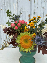 Close up of a small floral arrangement made in a turquoise ceramic pot. The flowers are in muted and bright fall tones, and the arrangement sits atop a white wooden table with a grey wooden background.