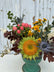Close up of a small floral arrangement made in a turquoise ceramic pot. The flowers are in muted and bright fall tones, and the arrangement sits atop a white wooden table with a grey wooden background.