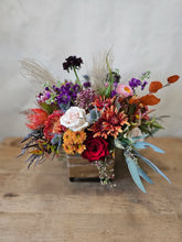 A floral arrangement made in moody fall tones, in a cube vase with tan ribbon tied around it. The arrangement sits on a wooden table. 