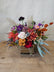 A floral arrangement made in moody fall tones, in a cube vase with tan ribbon tied around it. The arrangement sits on a wooden table. 