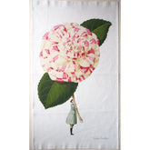 A white tea towel with an illustration of a woman in a green coat holding an oversized white and pink striped Camilla flower.