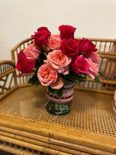 An arrangement of hot pink and light peach roses arranged in a clear glass vase with a red and white lace-inspired ribbon tied around it, sitting atop a wicker chair.  