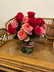 An arrangement of hot pink and light peach roses arranged in a clear glass vase with a red and white lace-inspired ribbon tied around it, sitting atop a wicker chair.  