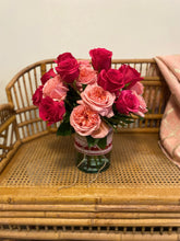 An arrangement of hot pink and light peach roses arranged in a clear glass vase with a red and white lace-inspired ribbon tied around it, sitting atop a wicker chair.  