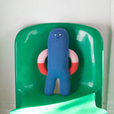 Blue plush toy with red and light pink arms on a green chair against a white wall.