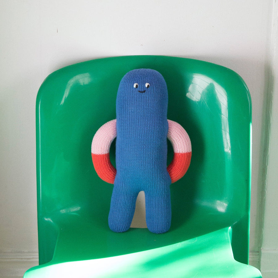 Blue plush toy with red and light pink arms on a green chair against a white wall.