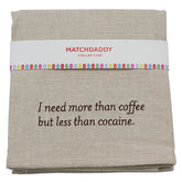 A folded beige linen tea towel with brown embroidered text reading 'I need more than coffee but less than cocaine.' and a white paper band labeled 'MATCHDADDY COLLECTION' with a colorful dotted border.