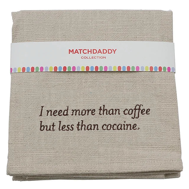 A folded beige linen tea towel with brown embroidered text reading 'I need more than coffee but less than cocaine.' and a white paper band labeled 'MATCHDADDY COLLECTION' with a colorful dotted border.