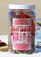 A clear glass jar filled with dried botanicals, topped with a white lid and a white-and-red label reading 'Day Drinking Daiquiri Craft Cocktail' and 'HB Craft Co.' A glass of mixed drink is partially visible around it on a white surface against a warm beige background.