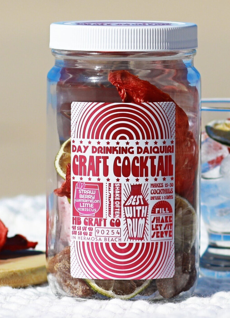 A clear glass jar filled with dried botanicals, topped with a white lid and a white-and-red label reading 'Day Drinking Daiquiri Craft Cocktail' and 'HB Craft Co.' A glass of mixed drink is partially visible around it on a white surface against a warm beige background.