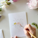 Person painting on a watercolor greeting card, with flowers surrounding the image slightly out of frame.