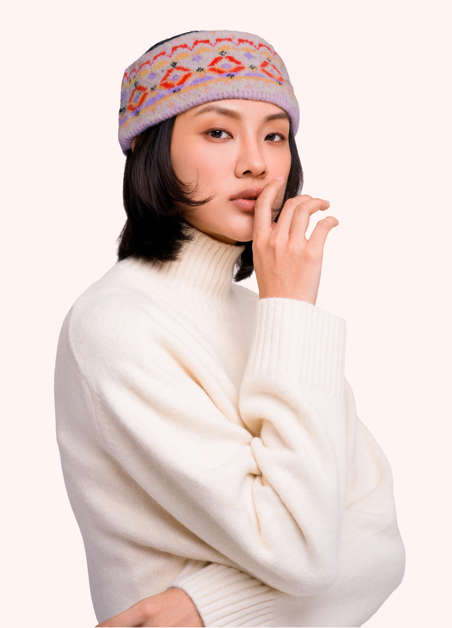 Woman in a cream colored sweater wearing a headband with a geometric orange, purple and gold pattern on it, against a light background.