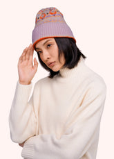 A woman in a cream colored sweater, wearing a beanie with a geometric orange, purple and gold pattern on it, against a light background.