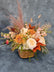 A medium sized floral arrangement in peach, pink, white and orange colors, arranged in a cube vase with brown and gold polka-dotted ribbon. The arrangement is set against a grey tie-dye background.