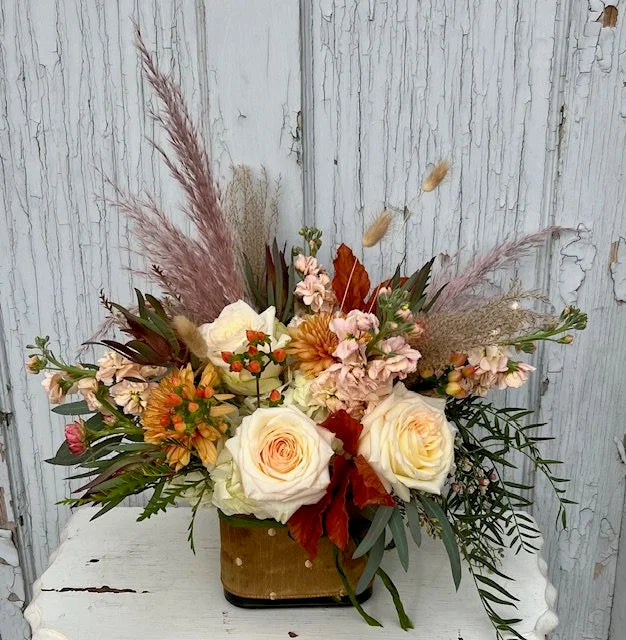 A floral arrangement in white, orange, pink, peach, and moodier colored flowers in a cube vase with a tan and gold polka-dot ribbon tied around it. The arrangement sits on a white wooden table with a grey wooden background. 