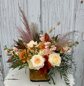 A floral arrangement in white, orange, pink, peach, and moodier colored flowers in a cube vase with a tan and gold polka-dot ribbon tied around it. The arrangement sits on a white wooden table with a grey wooden background. 