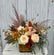 A floral arrangement in white, orange, pink, peach, and moodier colored flowers in a cube vase with a tan and gold polka-dot ribbon tied around it. The arrangement sits on a white wooden table with a grey wooden background. 