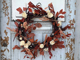 Grapevine wreath decorated with beech leaves, white wood flowers, bunny tail, and buttons. The wreath hangs against a grey wooden door. 