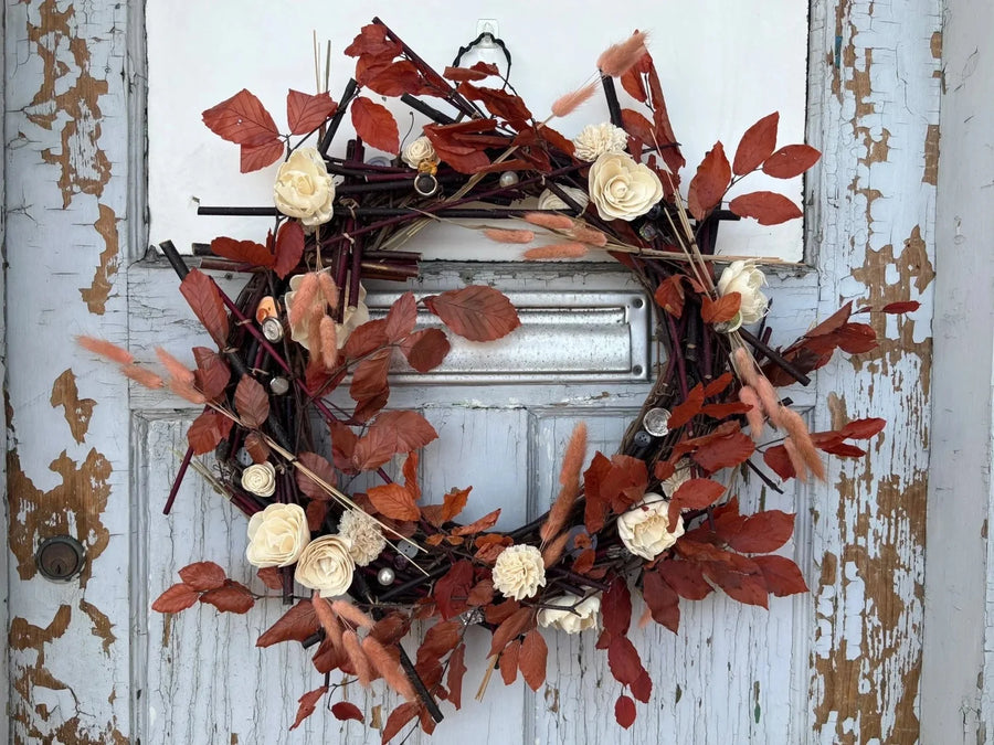 Grapevine wreath decorated with beech leaves, white wood flowers, bunny tail, and buttons. The wreath hangs against a grey wooden door. 
