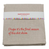 A folded beige linen tea towel with red embroidered text reading 'I hope it's the final season of this shit show.' and a white paper band labeled 'MATCHDADDY COLLECTION' with a colorful dotted border.