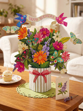 A large, paper bouquet with butterflies, colorful florals and a banner that reads "HAPPY BIRTHDAY" in a striped vase, sitting on a wooden coffee table with a matching card sitting to its right, and a cupcake on a small plate to its left. 