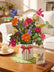 A large, paper bouquet with butterflies, colorful florals and a banner that reads "HAPPY BIRTHDAY" in a striped vase, sitting on a wooden coffee table with a matching card sitting to its right, and a cupcake on a small plate to its left. 