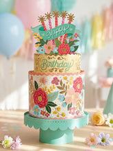 A large, paper pop-up card of a birthday cake decorated with florals and the words "HAPPY BIRTHDAY" written across it, on a cream table with pastel florals scattered across it. Balloons and other decor can be seen out of focus in the background.