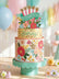 A large, paper pop-up card of a birthday cake decorated with florals and the words "HAPPY BIRTHDAY" written across it, on a cream table with pastel florals scattered across it. Balloons and other decor can be seen out of focus in the background.
