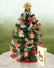 A large, paper Christmas tree pop-up card decorated with vintage-style ornaments such as rocking horses, angels, and doves. It sits atop a marble table with a matching card to its right.