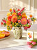 A colorful paper flower bouquet in a cream floral pattern vase, styled on a table with sliced citrus, pink tulips, and a matching card in a bright, warm interior setting. 