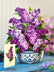 A large, paper bouquet of lilacs in a blue and white pattern vessel, with a matching card standing next to it on a blue wooden table. 